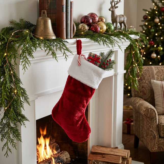 Red and white plush Christmas stocking hanging on a decorated fireplace mantel with garland and fire